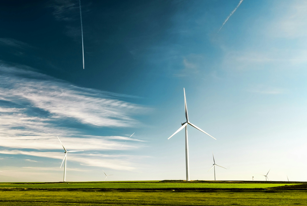 Green field with several wind turbines against a backdrop of a blue sky with streaks of clouds and jet contrails.